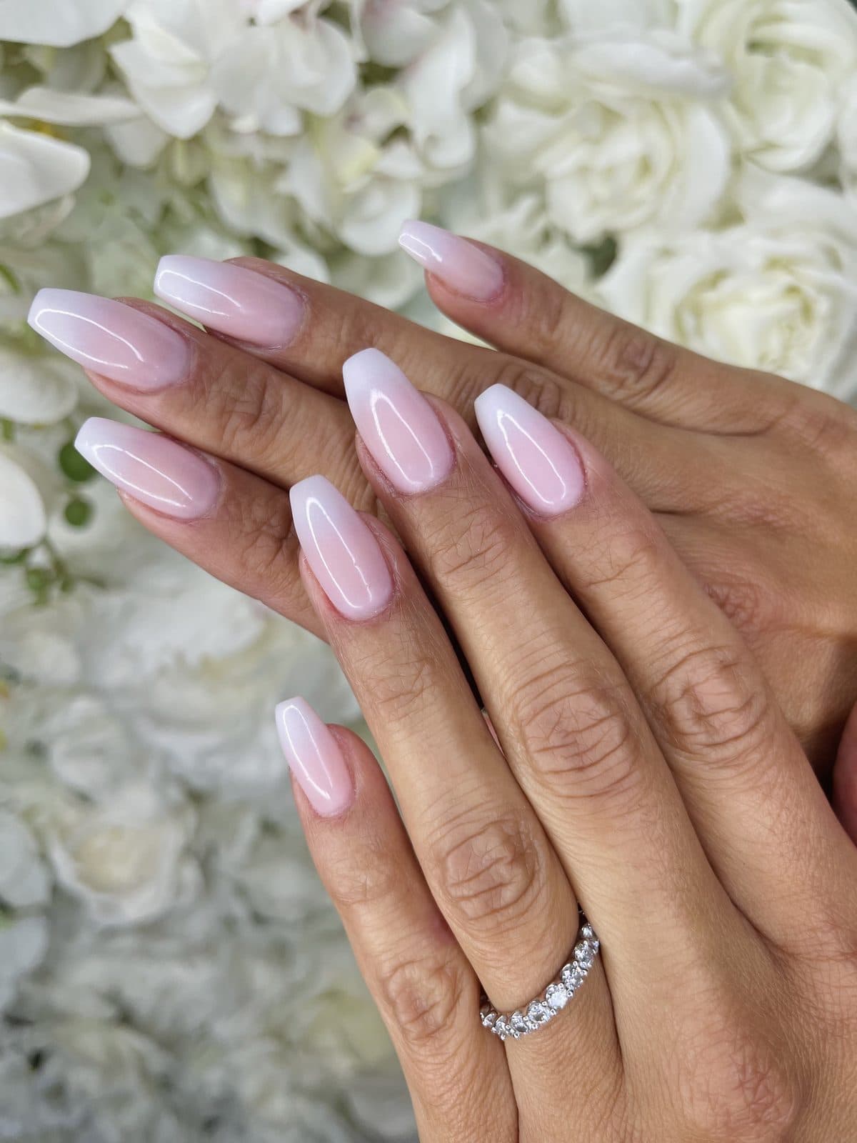 Des ongles en amande, d'un ombré rose pâle, devant le mur de fleurs